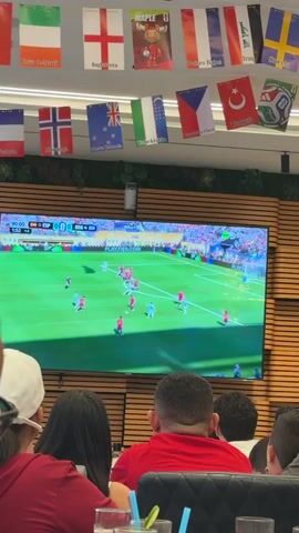 A ceiling-mounted television displays a live soccer match. Several people are visible in the foreground, watching the game from behind. The television is positioned above a wooden paneled wall. A string of international flags hangs from the ceiling above the television. The flags appear to represent various countries. The soccer match on the screen shows players in red and light blue uniforms on a green field. The scoreboard indicates the game is between ESP and ARG. Some of the spectators in the foreground are wearing red shirts. The people in the foreground are partially obscuring the bottom portion of the television screen. One person in the foreground is raising their hands, possibly in excitement or cheering. The environment appears to be indoors, possibly a sports bar or a similar venue. The lighting suggests an indoor setting. Drinks and glasses are visible on a table in the lower right corner of the frame.