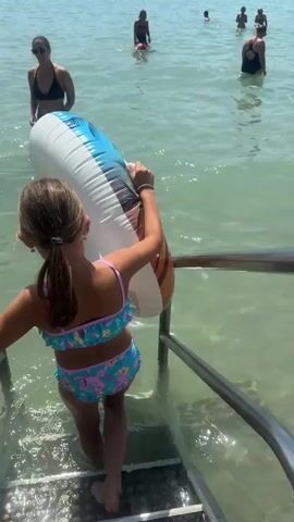 In this media, a young girl with her hair in a ponytail, wearing a colorful bikini, stands at the bottom of a metal staircase leading into clear, shallow water. She is holding a large, inflatable ring with a tropical design. Behind her, another child in the water is partially visible. Further back, several adults and children are enjoying the water, with some swimming and others standing. The scene appears to be a beach or lakeside setting on a sunny day, with calm water and a hazy sky in the distance, suggesting a warm climate. The overall mood is one of leisure and family fun. The location is identified as Annecy, France. There is no visible text in the media.