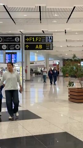 The image captures the interior of a modern airport terminal in Nouaceur, Morocco, characterized by a bright, spacious environment with a high, paneled ceiling and glossy tiled floors reflecting the light.

In the foreground to the left, a woman wearing a light-colored hoodie and dark pants walks away from the camera, holding a mobile phone. A small child's leg is partially visible beside her. In the mid-ground, a man in a light blue shirt and jeans walks towards the camera. To his right, two women dressed in dark business suits walk together, appearing to be in conversation. Other blurred figures can be seen in the distance, indicating general airport activity.

Prominently displayed are several large black and yellow overhead signs providing directions and information. One sign on the left points to "Prayer room" (مصلى) and "Coffee" (مقهى), along with "Tobacco." Another sign indicates directions to "-> F1,F2" and "↑ AB" gates, also showing "Gate T1" (بوابة T1) and "Gate" (بوابة / 登机口) in Arabic, English, and Chinese characters. A flight information display screen (FIDS) is visible in the background, along with various duty-free or retail shops. A small green plant in a wooden planter adds a touch of nature to the right side of the frame. The bright lighting suggests it is daytime. People appear to be moving with purpose, typical of an airport environment, with no strong emotions discernible.