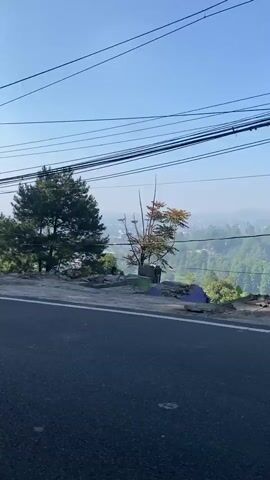 The video begins by showing a road and a tree with a background of a valley. Power lines are visible above. The camera then pans to the right, revealing a glimpse of a town in the valley below. Next, a white car and a truck carrying foliage are shown passing by. The video continues to pan right, showing more trees and vegetation along the roadside, with glimpses of houses and buildings in the background. The scene is set in a hilly or mountainous area with a clear blue sky.