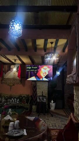 This image captures the interior of a traditionally decorated Moroccan cafe or lounge in Kenitra, Morocco, likely in the evening due to the artificial lighting.

**Subjects:** The scene features ornate Moroccan decor, including two large, decorative chandeliers – one with blue light on the left and a more intricate, darker one on the right. On the left wall, a framed portrait of a man, likely King Mohammed VI of Morocco, is displayed alongside two Moroccan flags. A large screen in the center of the room shows a woman's face, possibly a singer or actress. Below the screen, there's a microphone stand and speakers, suggesting a live music or karaoke setup. In the foreground, a low, round table is set with white cups, plates, and a book. The seating around the table consists of deeply cushioned sofas or benches covered in richly patterned fabrics. Potted plants add greenery to the right side of the room.

**Activity or event taking place:** The setup suggests a social gathering space, possibly a restaurant or cafe, where patrons can enjoy food, drinks, and entertainment, such as watching music videos or live performances.

**Notable details:** The decor strongly reflects Moroccan aesthetic with its intricate patterns, warm colors, and traditional lighting fixtures. The time of day appears to be evening or night, indicated by the sole reliance on artificial lighting. The overall atmosphere seems relaxed and inviting.

**Visible text:** On the screen, Arabic text is clearly visible. The top line reads "نجمه صباح" (Najmat Sabah - Star of Sabah), and below it, "فيريال كريم" (Ferial Karim), identifying the woman on screen. Further below, the phrase "كنت الظالم وخصمي" (You were the oppressor and my adversary) is displayed, likely part of a song title or lyrics. On the book on the table, some Arabic text is present, but it's not clearly decipherable.