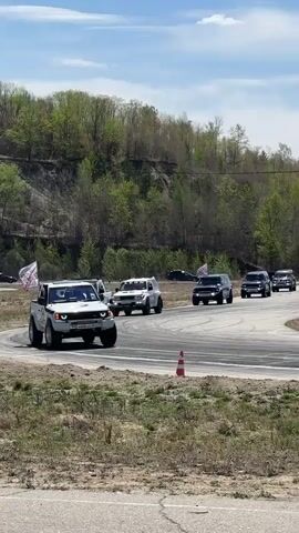 A group of off-road vehicles are seen driving along a paved course. The vehicles, a mix of white and dark colors, are mostly SUVs. Some vehicles have flags attached. The course is surrounded by trees and vegetation with a hill in the background. The sky is partly cloudy. The camera is stationary.