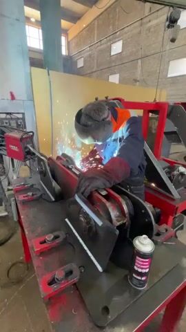 A welder in an orange-and-blue jumpsuit and black gloves is welding a red metal structure on a metal table. He is wearing a welding helmet to protect his eyes from the bright sparks. The welding machine is a red Lincoln Electric model. The surrounding environment is a large, industrial building with brick walls and high ceilings. There are windows providing natural light. Other tools and equipment are visible in the background, suggesting a workshop setting.