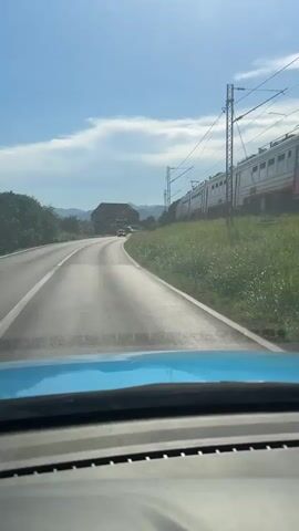 The video is filmed from inside a car looking forward through the windshield. A blue dashboard is visible in the foreground. The car is on a paved road that curves to the left. On the right side of the road, a train with red and white cars is moving parallel to the road on an elevated track. The train tracks are lined with power lines and poles. A grassy embankment with green vegetation runs alongside the train tracks. On the left side of the road, there are trees and some buildings in the distance. The sky is blue with scattered white clouds. The overall scene appears to be a sunny day in a rural or suburban area.