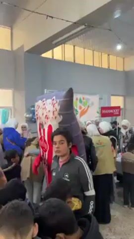 Here is a concise description of the video:

The video shows a gathering of people, mostly children and women wearing hijabs, indoors, possibly in a community or event hall. A person in an inflatable popcorn costume is surrounded by the children, who are looking at it with interest. Banners and logos are visible in the background, indicating a possible charitable event or organization. The atmosphere seems lively and cheerful.