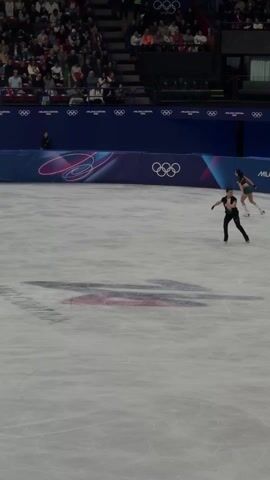 Two figure skaters, one female and one male, are performing on an ice rink in front of a large audience. The female skater is wearing a turquoise and red outfit, while the male skater is wearing black attire. The backdrop features the "MILANO CORTINA 2026" logo and Olympic rings. The ice rink has markings and logos, including a faded emblem in the center. Numerous photographers and videographers are positioned along the barrier. The audience fills the stadium seating, with flags visible in the crowd.