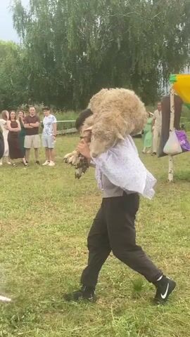 A man wearing a white shirt and black pants carries a sheep on his shoulders. He is surrounded by people in an outdoor setting with trees and grass. Some people are sitting on chairs, while others are standing and watching. A few people are holding up phones to take pictures or record videos. The man is walking and appears to be part of an event or performance. The sheep is light-colored and has a woolly coat. Some of the spectators are clapping. The scene suggests a festive or celebratory occasion.