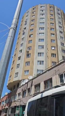 This is a vertical video of a tall, circular residential building in Ataşehir, Turkey. The building has a beige and light gray facade with numerous windows and external air conditioning units. A spiral staircase is visible on one side of the building, running from near the top to a lower level. In the foreground, a lower, older building with a brown facade and several windows is partially visible. A white vehicle, likely an SUV, is parked in front of the lower building, with its front section filling the bottom of the frame. The sky is clear and blue, suggesting a sunny day. The overall impression is of an urban environment with a mix of modern and older architecture. No people are clearly visible. The time of day appears to be daytime due to the bright sunlight.