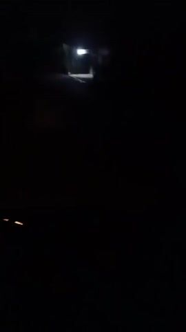 The video begins with a dark, blurry shot of what appears to be a dashboard or interior of a vehicle, with a circular light visible on the left side. The camera then pans to the right, showing a dimly lit outdoor scene at night. A paved road or path is visible, leading towards a structure with a bright light illuminating its entrance. The structure appears to be a small building, possibly a shop or house, with windows and a covered entrance. Vegetation can be seen on the left side of the frame. The camera continues to move, offering brief glimpses of the building and the surrounding dark environment. The overall impression is of a night-time drive or walk past a dimly lit building.