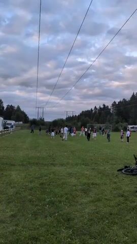 A group of people, primarily children, are playing soccer on a grassy field. The sky is cloudy. There are power lines running across the top of the frame. In the background, there is a line of trees and some buildings, possibly campers or trailers. A bicycle is lying on the ground in the foreground.