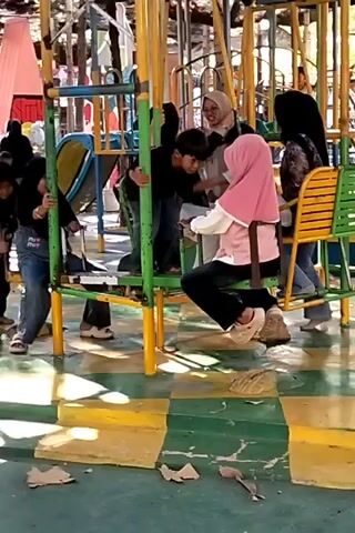 A group of children and adults are at a playground. In the foreground, a girl in a pink hijab and a white shirt is sitting on a swing, looking at the camera. To her left, a boy in a black shirt is on another swing, and a girl in a blue denim jacket and black pants is standing next to him. Further to the left, another girl in a black hijab and jeans is standing near a climbing structure. Several adults are visible in the background, some of whom appear to be supervising the children. The playground equipment is made of metal and painted in bright colors, including yellow and green. The ground is paved with checkered tiles in yellow and green. The scene is outdoors, with trees and foliage visible in the background. The lighting suggests it is daytime.