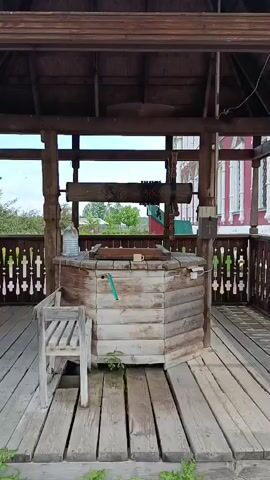 A wooden well with a roof is shown in detail. The well is constructed of logs and has a wooden lid. A chain mechanism with wooden handles is used to lower and raise a bucket. A white plastic bottle, a yellow mug, and a green hose are on top of the well. A wooden chair is placed beside the well. The well is located on a wooden deck with decorative railings. In the background, there are trees and a building with a red facade. The sky is visible through the roof. The video is a close-up of the well and its surroundings.