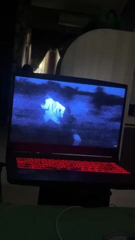 The video shows a laptop with a red-lit keyboard. The laptop screen displays a blue-toned image of a person, possibly wearing a white garment, standing in a dark, outdoor environment. The person appears to be the central focus of the image on the screen. The background of the image is dark and indistinct. The overall lighting of the scene is dim, suggesting it is either nighttime or an indoor setting with low light.
