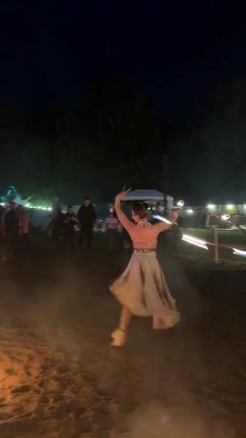 A woman in a cream-colored dress performs a fire dance with a stick that has fire on both ends. She is surrounded by a large crowd of people standing in a grassy area with trees, tents, and bright lights in the background. The ground she dances on is sand and there is a fire pit in the bottom left corner.