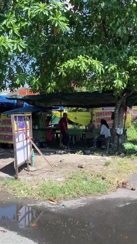 The video shows an outdoor food stall under a large tree. Several people are present. In the foreground, a paved road with puddles is visible. A grassy area with some fallen coconuts is to the right. A white board with writing is propped up on the left. The food stall has a green counter and a yellow awning. Signs on the stall read "LONTONG SAYUR" and "SOSEK". Two people are seated at the stall, and another person is standing near the counter, interacting with someone behind it. The environment appears to be sunny with shadows from the tree.