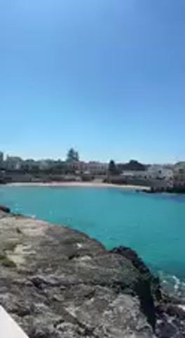 This video features a scenic panning shot of a coastline under a clear, bright blue sky. The camera initially shows a small bay with turquoise water, a sandy beach, and buildings of a distant town. As the camera pans right, it reveals a wider expanse of the vibrant turquoise sea, which is remarkably clear, allowing visibility of rocks beneath the surface. The foreground consists of dark, rugged rocky shorelines and a light-colored paved area, possibly a walkway or dock. The water transitions from a lighter turquoise near the shore to a deeper blue further out, meeting a distinct horizon line. The weather is sunny and calm, with no clouds visible. The scene depicts a beautiful coastal area in Nardò, Italy, enjoying a bright day.