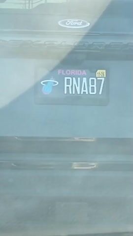 The video depicts a daytime traffic scene on a multi-lane road, likely in Cutler Bay, Florida, under a clear, sunny sky. The initial close-up focuses on the rear of a black Ford Explorer SUV. Visible on the tailgate are the "Ford" logo and partially "EXPLORER" lettering.

The SUV features a Florida license plate that prominently displays the Miami Heat basketball team logo. The plate number is "RNA87", and a small yellow sticker indicates "11-26".

As the camera zooms out, it reveals the broader context of a traffic jam, with multiple vehicles stationary or moving slowly. The Ford Explorer's brake lights are illuminated. To the left, a silver Chevrolet Silverado pickup truck is visible, also caught in traffic. The Silverado's rear window displays several lines of text, including a phone number "305-668-3335", which corresponds to the Miami-Dade county area code, further confirming the location in South Florida. Overhead traffic signal structures are visible against the bright sky. Background music is audible, indicating it's playing from the recording vehicle.