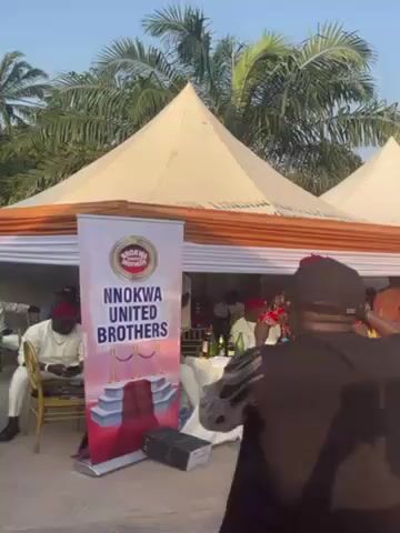 The video shows a group of people at an outdoor event. The event appears to be organized by "NNokwa United Brothers", as indicated on a banner. Men dressed in traditional attire are greeting each other. Some are seated while others are standing. The event takes place under white tents, and palm trees are visible in the background. Someone is speaking into a microphone.