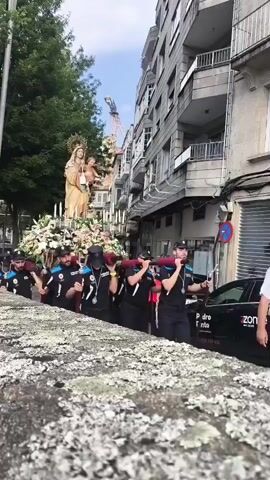 A group of uniformed men in dark blue shirts with light blue accents on the collar and sleeves are carrying a large, ornate float adorned with flowers. The float has a statue of a woman holding a child, believed to be religious figures. The men are walking in procession down a street lined with buildings and trees. Some of the men are wearing black baseball caps. One man in the foreground is wearing sunglasses and a necklace. The procession appears to be taking place outdoors during the day.