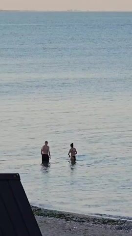 Two people, a man and a woman, are wading in the calm sea near a pebble beach. The man, facing away from the camera, is wearing dark swim trunks and is submerged up to his mid-thighs. The woman, also facing away from the camera, is wearing a bikini and is submerged up to her waist. She appears to be gesturing with her right arm towards the man. The water is a serene blue-grey, and the sky above is a pale, hazy orange, suggesting either sunrise or sunset. In the distance, a faint, hazy shoreline with structures is visible. The foreground is dominated by a dark, angled object, possibly part of a structure or a boat, with a hint of green vegetation and pebbles on the beach. The overall mood is tranquil and serene. There is no visible text in the media. The location is identified as Shabla, Bulgaria.