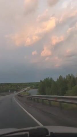 The video shows a car traveling on an asphalt road with a metal guardrail beside it. A body of water is visible on the other side of the guardrail, with trees lining the opposite shore. The sky is cloudy with pink and gray hues. The camera appears to be mounted inside the car, looking through the windshield.
