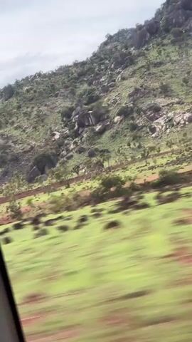 The video shows a view from inside a moving vehicle, looking out at a rural landscape. A green, rocky hillside covered in trees and shrubs dominates the background under an overcast sky. In the foreground and mid-ground, there are fields with reddish-brown soil and patches of green vegetation, suggesting cultivated land or natural grasslands. The blurriness of the immediate foreground indicates the vehicle's motion. The scene depicts a journey through a natural, undeveloped area with some agricultural elements, likely in Gidan Waya, Nigeria, given the context.