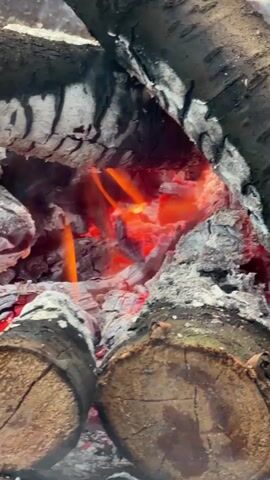 A close-up shot shows a campfire with glowing red embers and flames within a structure of burning logs. The logs are charred and split, with white ash scattered around them. The fire flickers and shifts, producing a warm orange glow that illuminates the scene.