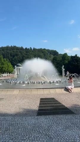 The video shows a large, ornate fountain with a central spire and multiple smaller jets of water spraying upwards. The fountain is surrounded by a shallow pool filled with metallic spheres, and a paved area with a cobblestone pattern. A woman in a white dress is kneeling by the edge of the fountain, taking a picture with her phone. In the background, there is a colonnade with a dome, lush green trees, and a dense forest on a hillside under a clear blue sky. The lighting suggests it is a sunny day, likely late morning or early afternoon. The overall atmosphere appears serene and picturesque, a common scene in a spa town like Mariánské Lázně, Czechia, known for its mineral springs and elegant architecture. The woman's posture and focus on her phone indicate she is capturing a moment of enjoyment or documenting her visit.
