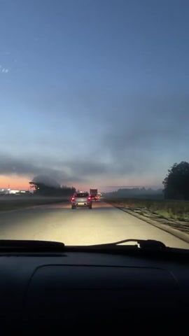 The video is filmed from the interior of a car, looking forward. The car is on a paved road with lane markings visible. The sky is a gradient of blue and orange, indicating either dawn or dusk. Clouds are present in the sky. In the distance, there are several vehicles on the road, including a car with its headlights on and a truck. There are also lights visible on the sides of the road, suggesting buildings or infrastructure. The landscape on either side of the road appears to be fields. The overall impression is a drive during twilight.