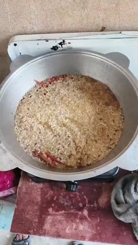 A top-down shot shows a large metal pot on a stove. The pot is filled with rice and some pieces of red meat or vegetables. The contents of the pot are bubbling, indicating that they are cooking. The pot is round and has handles on either side. The stove appears to be electric, with a flat, white surface. There is a wooden surface beneath the stove, with some pink and red objects on the left side of the frame and a grey cloth on the right. The background is a light-colored, textured wall.