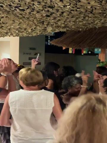 The video captures a lively outdoor gathering, likely at night, given the artificial lighting and the dark background that suggests evening or night time. The setting appears to be a casual, festive venue, possibly a beach bar or outdoor lounge area, given the thatched roof overhead and string lights, some of which are displaying colors. Small flags of various countries are hung across the scene, suggesting an international or celebratory atmosphere.

The central activity is a group of people dancing and socializing. In the foreground, several individuals are visible from behind, some with their backs to the camera, suggesting they are facing the dance floor or a stage. Their attire is casual, with one person wearing a white t-shirt with a colorful graphic design on the back. Further into the scene, more people are engaged in dancing and interacting. One individual in the middle appears to be holding up a phone, possibly recording the event. There's a person in an orange shirt and another with a colorful headdress, contributing to a festive mood. The overall impression is one of enjoyment and merriment.

There is no visible text in the video that provides specific information about the event or location, beyond the country flags hinting at an international theme. The emotions conveyed are predominantly positive, with people appearing to be having a good time. The weather is not explicitly discernible, but the outdoor setting and the presence of many people suggest it is pleasant. The time of day is clearly evening or night due to the lighting conditions.