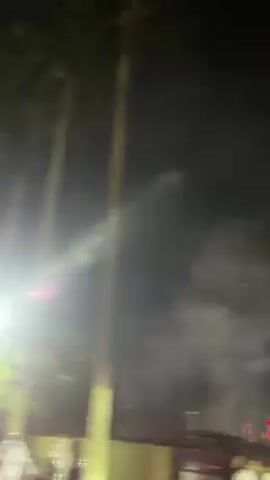 The video shows fireworks exploding in the night sky. In the foreground are tall palm trees silhouetted against the darkness. There's smoke in the air, possibly from the fireworks. A street light provides some illumination, and the roofs of nearby buildings are visible. The fireworks themselves are bright and colorful, bursting into a variety of patterns in the sky. The camera angle is from below, looking up at the fireworks.