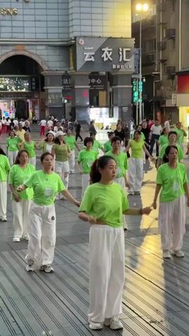 A group of women are performing a coordinated dance routine in a public square. They are uniformly dressed in light green t-shirts and white pants, creating a sense of unity. The group is of varying ages. The dance is synchronized, with the women moving in unison. The setting is an urban environment, with buildings visible in the background. Other people, some of whom are passersby, are present in the background. The performance takes place on a paved surface.