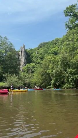 The camera is still while a body of water is observed, with canoes lined up along a bank of the river. The water is slightly rippled with a brown tint. A small cluster of people are on the riverbank near the canoes. The trees surrounding the river bank are lush and green. In the background is a large rocky cliff face reaching high into the blue sky. The weather appears to be sunny and clear.