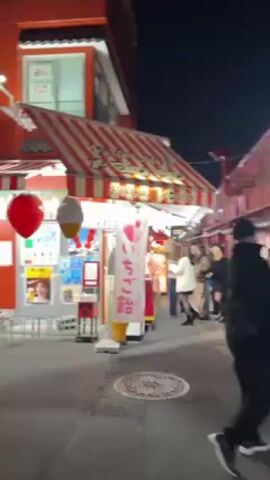 A short video shows a street food stall in Japan at night. The stall has a red and white striped awning with Japanese writing and advertisements for melon bread and strawberry candy. Several people are standing in line to order from the stall. A woman wearing a white jacket is standing at the counter, looking at the candy. The stall has glass displays filled with colorful strawberry candy. The street is narrow, and the buildings on either side are painted red. There is a sewer grate in the foreground. The overall atmosphere is lively and festive.