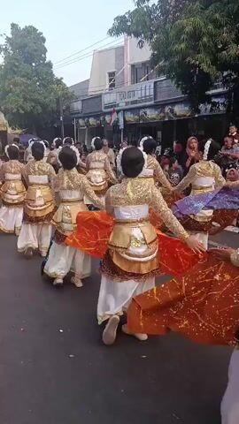 A group of dancers in traditional ornate gold and white costumes with colorful sashes are performing a choreographed routine in the street. They are arranged in a line, moving forward and to the right from the camera's perspective. The dancers have their hair styled up with white floral accents. The background shows a street with buildings, trees, and a clear sky. A red and black balloon, possibly shaped like Spiderman, is visible on the left side of the frame. The dancers' movements create a flowing effect with their sashes, which are orange and green in the foreground. The overall scene appears to be a procession or festival.