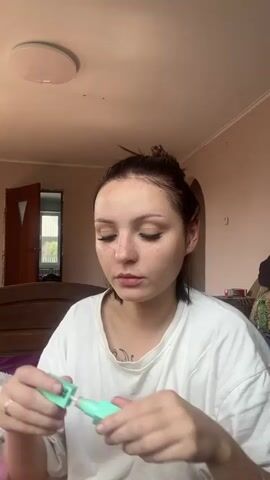 A young woman with dark hair pulled back into a bun and wearing a white t-shirt applies concealer to her face. She holds a tube of concealer in her left hand and the applicator in her right, dabbing the product under her eyes and on her cheeks. The background shows a room with pink walls, a door, and a bed.