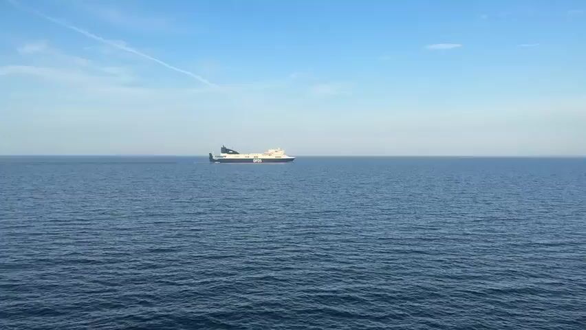 A cargo ship sails across the deep blue sea under a bright, partly cloudy sky. The ship is white and dark blue with the letters DFDS visible on its side. The water is calm with small waves. The horizon is visible in the distance, showing a faint line of land. A faint white contrail is visible in the sky.