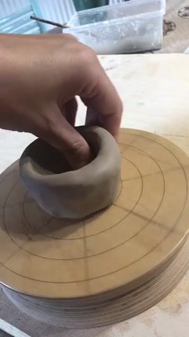 A close-up shot shows a person's hand shaping a lump of clay on a wooden pottery wheel. The clay is a light brown color and has been formed into a roughly bowl-like shape. The person's thumb is inserted into the center of the clay, pushing it outwards and downwards to create the interior of the bowl. The pottery wheel has concentric circles and radial lines marked on its surface, presumably for guidance during the pottery-making process. In the background, a white plastic container with some material inside, a green mug, and a wooden surface are visible. The lighting suggests an indoor setting.