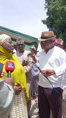 The image captures an outdoor interview or press briefing taking place in Ungwan Liman, Nigeria, under bright daylight with a clear blue sky.

In the foreground, a woman, likely a public figure, is speaking into a red microphone bearing the "CHANNELS" logo. She wears a light-colored headwrap, a vibrant yellow patterned shawl, and a white patterned dress. Her mouth is open as if mid-sentence, and her expression appears serious and engaged.

Opposite her, a man in a white embroidered long-sleeved shirt, dark red and black striped trousers, a brown straw fedora hat, and sunglasses holds a blue microphone with the "VON" (Voice of Nigeria) logo. He is looking at the woman, listening intently.

Behind the primary speakers, several other individuals are present, observing the event. One man behind the woman wears a light brown traditional outfit and a patterned cap, holding a drink and a mobile phone. Other bystanders, some in traditional attire, are also visible, with several holding up phones, suggesting they are recording or taking photos.

The setting includes green foliage from trees or bushes in the mid-ground and a light-colored building with a darker roof in the background. The ground beneath the participants appears to be loose earth or gravel. The bright lighting indicates it is likely midday or early afternoon. The overall scene suggests a formal interaction with the media.