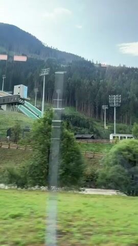 The video shows a ski jump facility located on a forested mountainside. The ski jump ramp is visible, with lights surrounding it. The surrounding area is covered in trees, and the sky is cloudy. The camera appears to be moving, likely from a vehicle, as evidenced by the motion blur in the foreground. The scene is natural and mountainous, with the ski jump being the man-made structure within it.