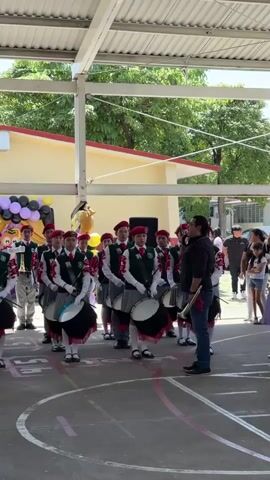 A group of young people in matching uniforms, consisting of red berets, white shirts, green vests, and plaid skirts or trousers, are lined up in a formation outdoors under a covered structure. They are holding drums and appear to be part of a marching band or parade. A man wearing a dark shirt and jeans stands in front of them, facing them and holding a baton or stick, seemingly directing them. In the background, there is a building with a yellow facade and a red-tiled roof. Balloons in black, purple, and yellow are visible, as is a yellow inflatable character, suggesting a celebratory event. Other people are standing or walking in the background. The ground is paved with white and pink markings, resembling a sports court. The lighting suggests it is daytime, with natural light illuminating the scene.