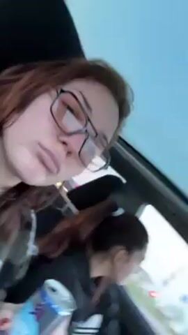 A woman in glasses is in the passenger seat of a car, apparently filming a selfie video. She is sticking her tongue out. Another woman is in the driver's seat with her hair pulled back. The car is in motion. The sky is overcast. The woman in the passenger seat has a can of soda in her hand. Loud music plays in the background.