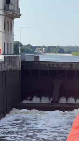 A view of a dam or lock gate with water rushing through. The water is turbulent and foamy. There is a large concrete structure to the left with a balcony. In the background, there is a body of water with trees and a building on the far shore. An orange object, possibly a life raft or buoy, is in the foreground to the right. The sky is clear and blue.