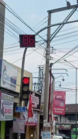 The video shows a street scene with a traffic light that is red. A digital display above the traffic light shows the numbers counting down from 86 to 82. The display is attached to a pole that also carries many electrical wires. To the left of the traffic light, there is a building with several signs, including one that says "Cakrawala PHOTOCOPY & DIGITAL PRINTING". To the right, there are more signs and a pole with electrical equipment. The sky is visible and appears overcast. Several cars are visible in the background.