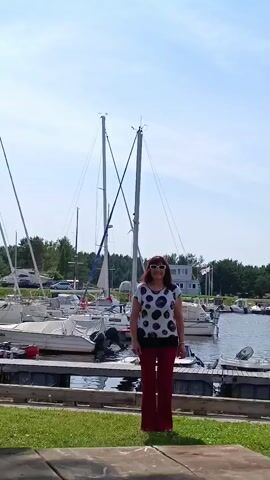 A woman stands on grass with her arms raised and palms facing forward. She is wearing a white shirt with black polka dots and red pants. Behind her, a marina is filled with sailboats. The sky is clear and blue.
