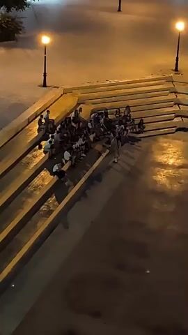 The video captures a group of people sitting on wide, concrete steps at night. Streetlights illuminate the scene, casting a warm glow on the square and the surrounding buildings. The individuals, appearing to be young adults, are gathered together, some talking and others looking around. The atmosphere seems relaxed and informal, suggesting a social gathering or a casual hangout. The architecture of the buildings is typical of an urban setting, with balconies and multiple floors. The ground is wet, reflecting the artificial lights, indicating that it may have recently rained.
