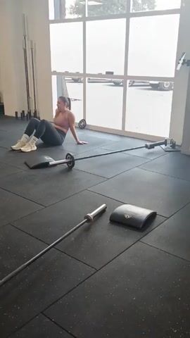 A young woman is sitting on the floor in a gym. She has dark hair pulled back in a ponytail and is wearing a mauve-colored sports bra and dark leggings. She is leaning back on one arm, with her knees bent and feet flat on the floor. In front of her are two barbells and two black pads. The floor is covered in black rubber mats. Behind her is a large window that looks out onto a parking lot. There are other gym equipment in the background.