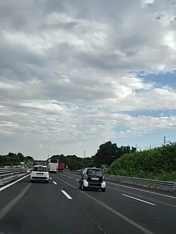 This is a first-person perspective video clip from inside a vehicle traveling on a highway. The time of day appears to be daytime, with a mix of sun and clouds in the sky. The weather seems fair.

The main subject of the clip is the highway itself, with multiple lanes of traffic. Several cars are visible, including a small dark blue and silver Smart car in the foreground, a white compact car further ahead, and a white bus. In the background, there is a red truck and other vehicles. The surrounding environment includes lush green trees and vegetation along the sides of the highway. A railway bridge with overhead electrical wires is visible in the distance. There is also a road sign with a green background and a white symbol, possibly indicating a service area or an exit.

The activity is simply traveling on the highway. The overall mood is neutral, suggesting a routine journey. There is some visible text on the license plate of the Smart car, which appears to read "CR 748ZL". There is also some text on the green road sign, but it is not clearly legible. The location context is Teano, Italy, which aligns with the presence of Italian license plates and the style of road signage.