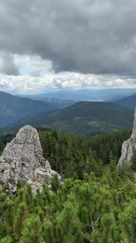 The video pans across a mountainous landscape. In the foreground, there are the tops of green pine trees. Behind them, a large, jagged rock formation stands prominently. Beyond the rock formation, a dense forest of dark green trees stretches out, forming hills. In the distance, more rolling hills and mountains are visible under a cloudy sky. The clouds are dark and appear heavy, suggesting potential rain. The overall scene depicts a rugged, natural environment.